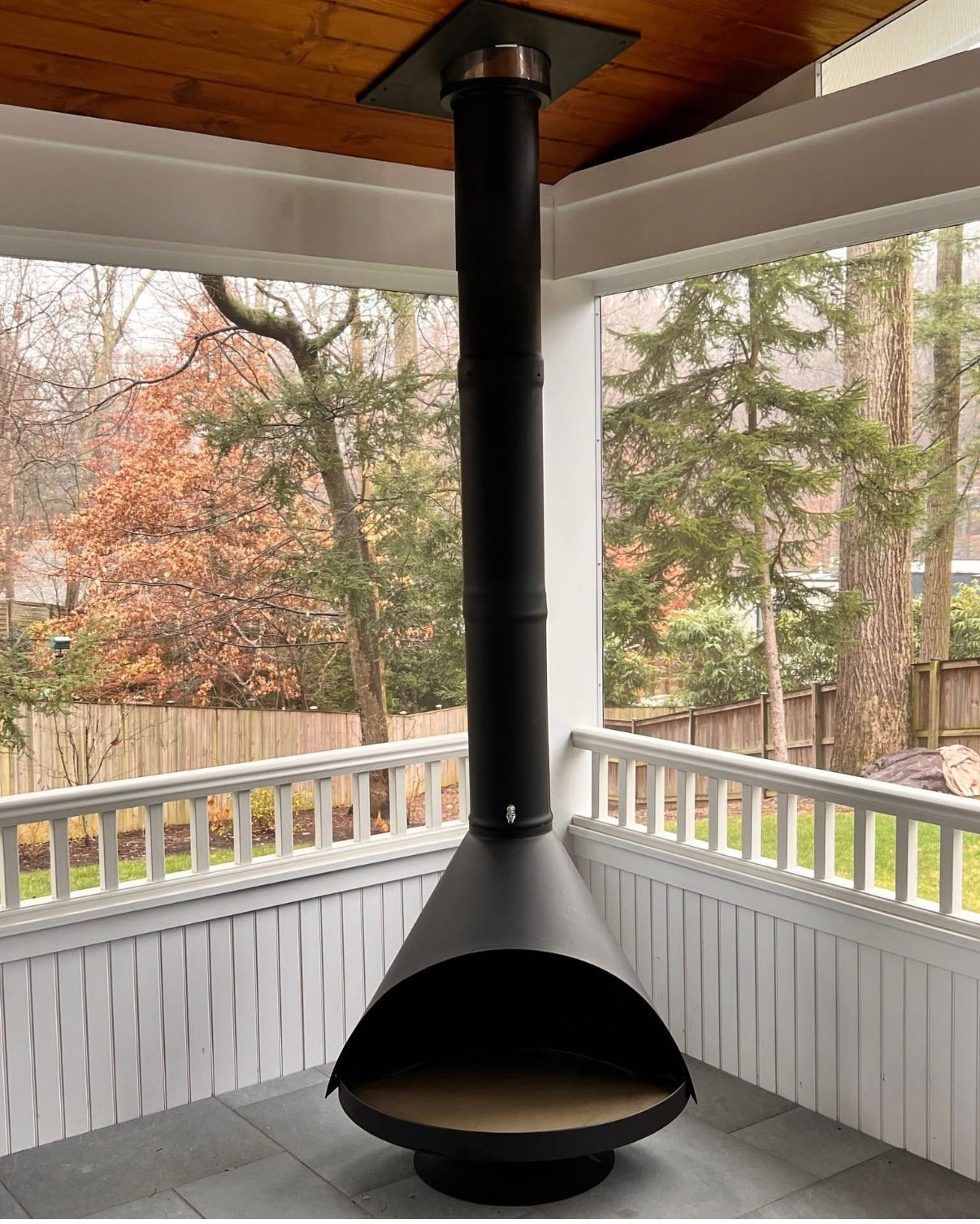 Black outdoor fireplace mounted on a wooden deck with a view of trees and a fence. Corner Fireplace on a covered patio
