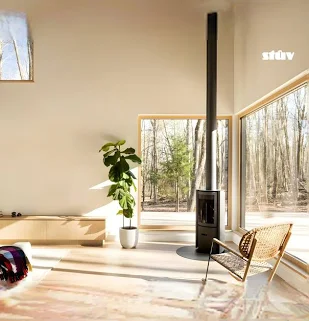 Stûv S30C compact wood burning stove installed in a minimalist modern home with floor-to-ceiling windows overlooking a forest — freestanding Belgian design stove