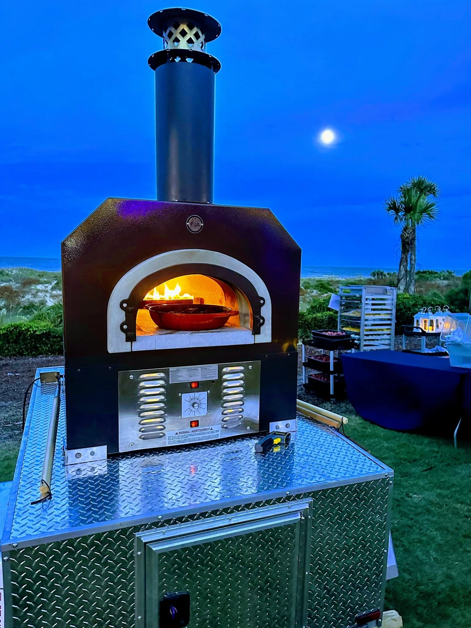 Outdoor view of CBO 750 Tailgater pizza oven with fire inside, set against a night sky with a moon.