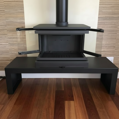 A modern black metal fireplace insert with a glass wind guard and a white chimney pipe, set on a wooden floor against a beige and wood-paneled wall.