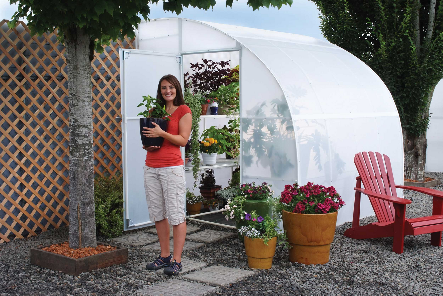A smiling on a red dress holding a black vase standing in front of 8' X 24' solexx harvester small greenhouse