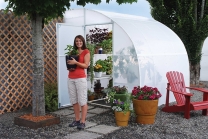 A smiling on a red dress holding a black vase standing in front of 8' X 24' solexx harvester small greenhouse