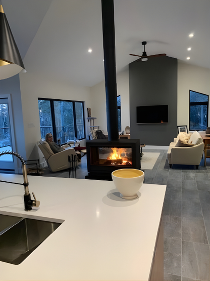 Invicta Aaron Double Sided 36" cast iron Stove on a small bench with Black Chimney connected to the room in a middle of modern living room.