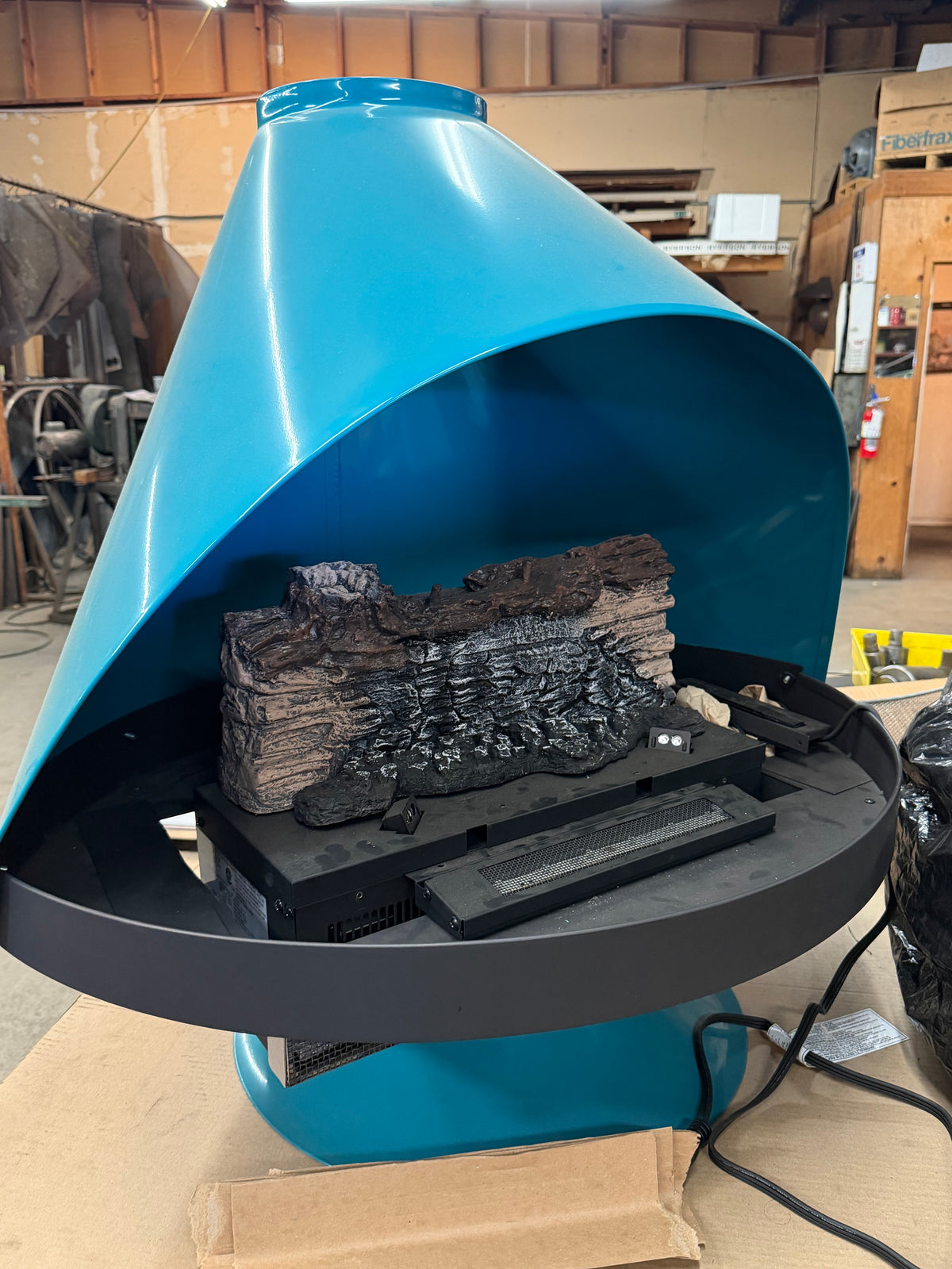 A teal blue electric fireplace insert with a black circular base and a realistic black and brown faux log set, photographed in a workshop environment.