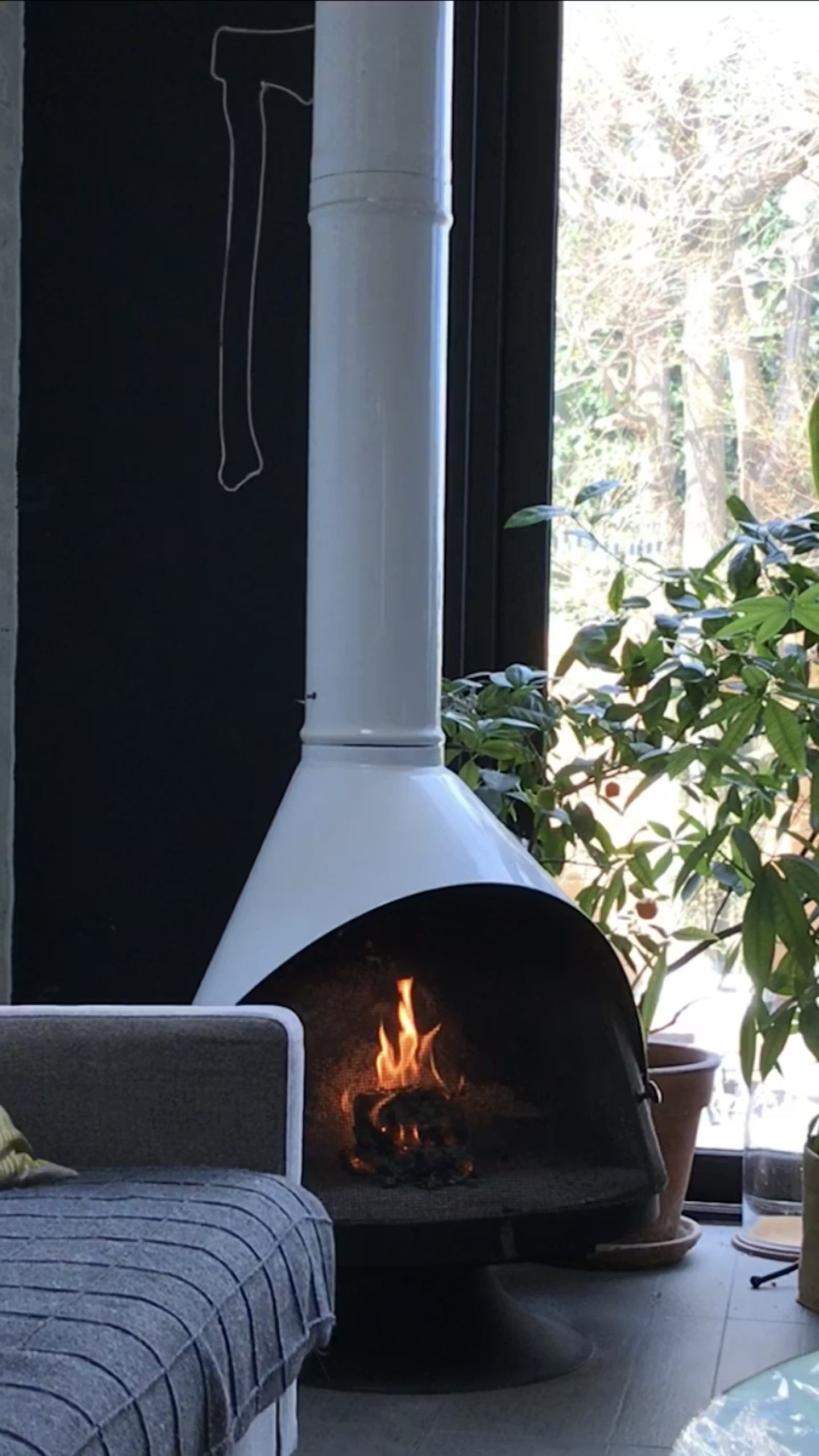 White coated Top Finish and Matte Black Base Finish Cone shape Malm 30" zircon wood burning fireplace with fire burning next to a gray sofa and a window with greenery outside