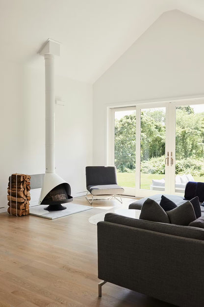 White Coated Top Finish and a Matte Black Base Finish Cone shape Malm 30" zircon wood burning fireplace in a Modern living room with sofa, and large windows.