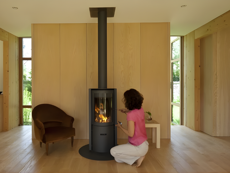 Woman in a pink shirt and white pants kneeling in front of a modern Stuv 30C wood burning stove in a room with wooden walls and floor.