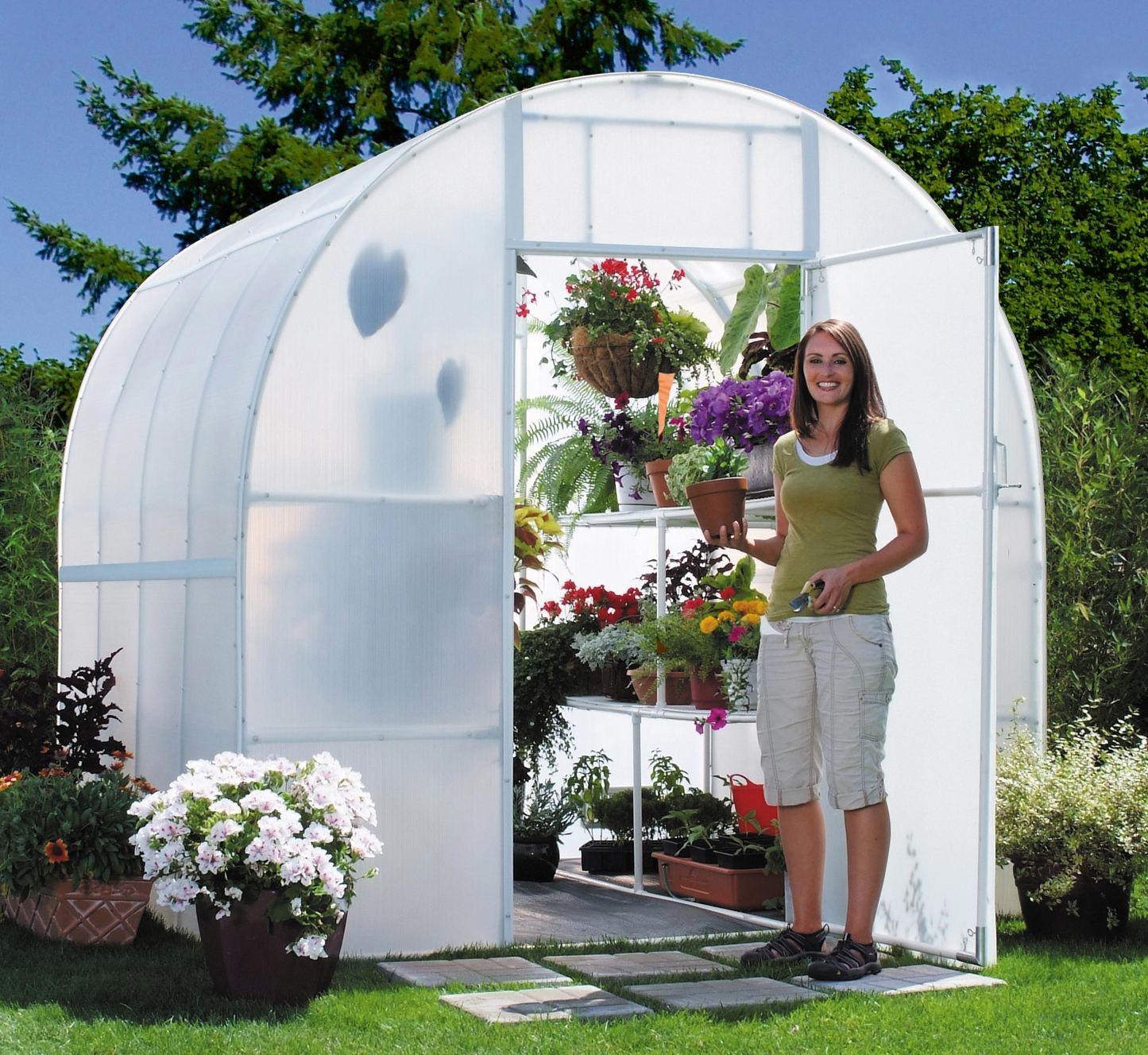 A woman standing holding small vase of green plant is standing in front of 8' X 8' Solexx gardener's Oasis Outdoor greenhouse