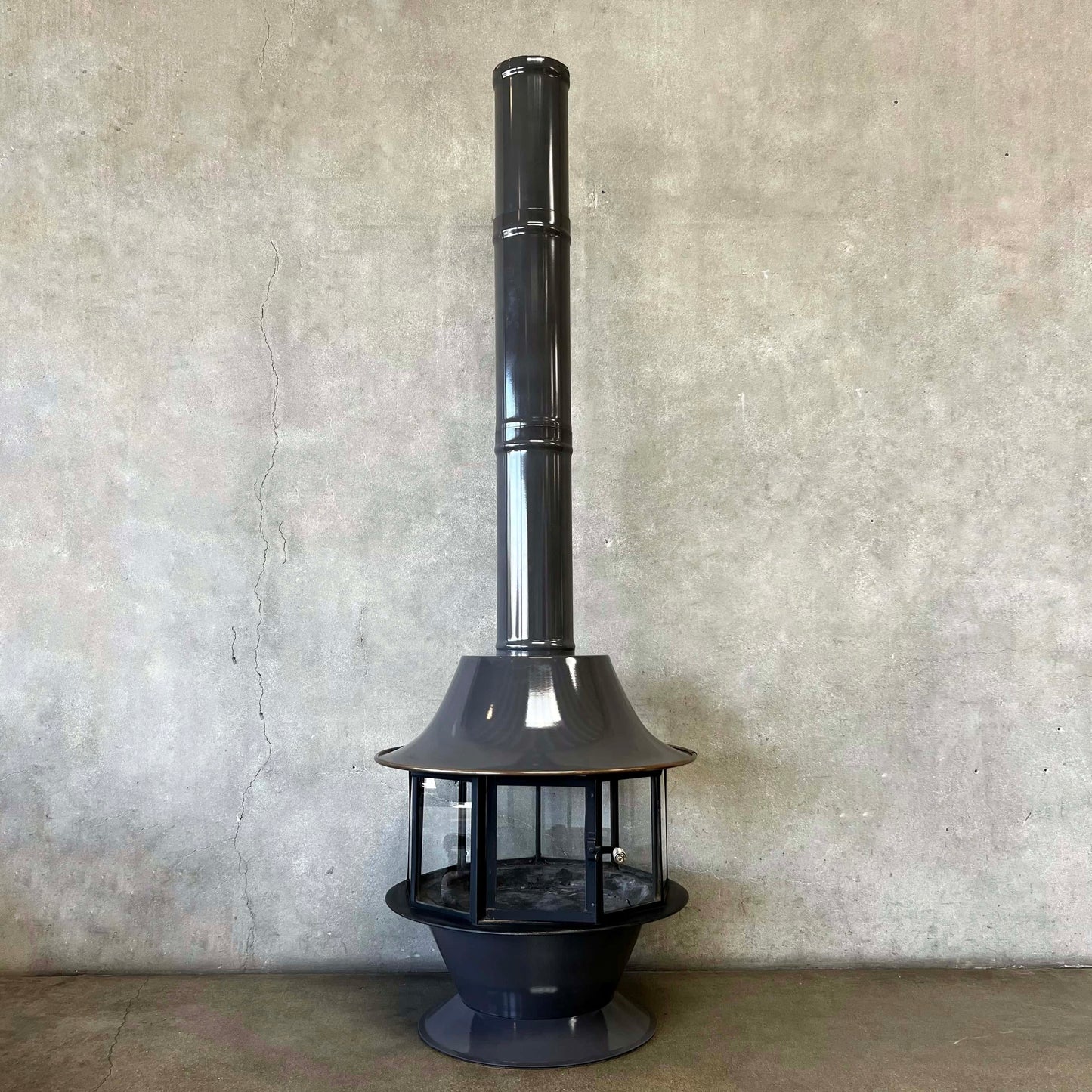 Black metal chiminea against a textured wall. throught he wall firepalce Slate Grey MCM Fireplace Spin a Fire custom color bright colored Carousel zircon, Focus Floating Fireplace hanging fireplace levitating fireplace Metallic outdoor light fixture on a concrete surface with a neutral background