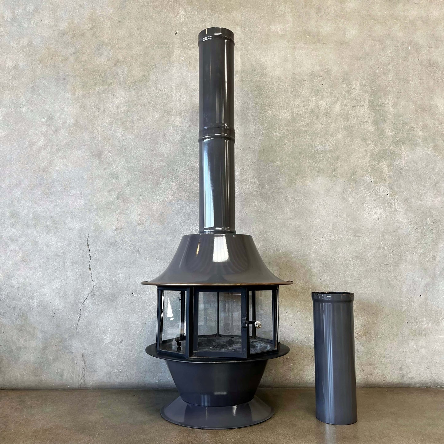 Black metal chiminea with a smaller cylindrical component against a textured wall.