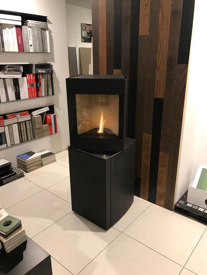 Pivot head dark oak base free standing Stuv S-P10 pellet stove with visible burning flame in a corner of a modern living room. Modern Indoor remote control and Smart phone control Home heating fireplace. Eco-Friendly, Gentle flame Indoor fireplace. This Fireplace is similar to ComfortBilt Alpine HP40 Pellet Stove, Osburn Volta Pellet Stove, Quadra-Fire Outfitter II Pellet Stove Display Demo,US Stove 1300 Sq ft Wood Pellet Stove, Castle | Serenity Pellet Stove.
