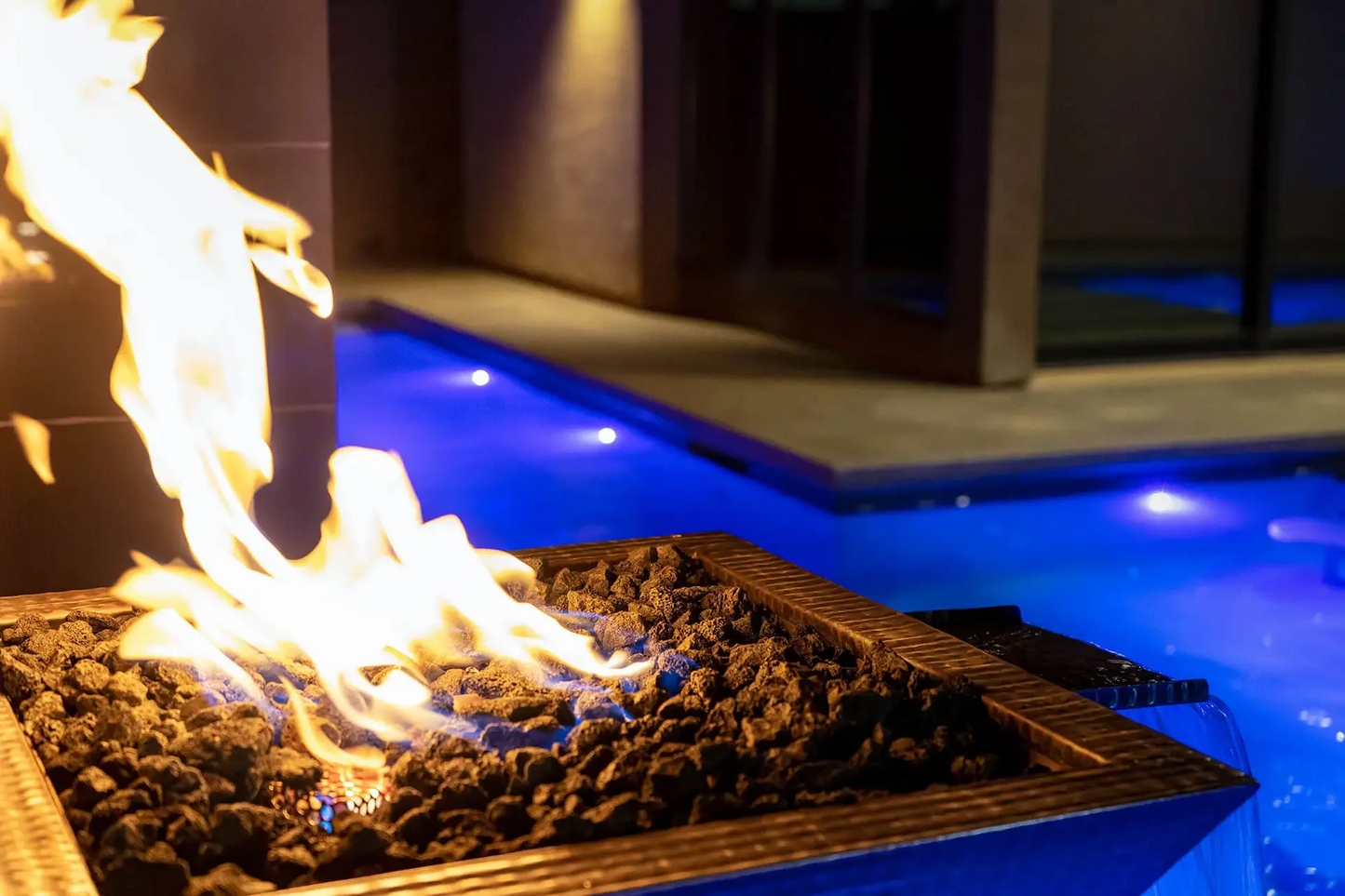 Close-up of a glowing fire feature with a luxurious pool illuminated in blue at night in the background.