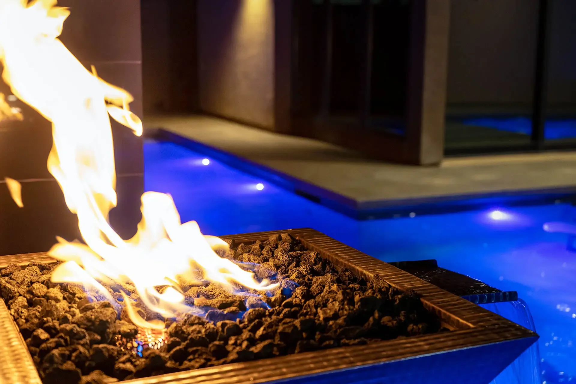 Close-up of a glowing fire feature with a luxurious pool illuminated in blue at night in the background.