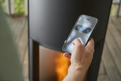 Hand holding a smart phone to control Matte Black Stûv P-20S Freestanding Modern Indoor pellet burning stove in large close spade with visible burning flame and pellet twin set container beside the fireplace. Elegance, silence, performance, ease of use and maintenance Fireplace. This pellet stove is similar to Stûv 30 Wood Stove | Compact Glass Fire Screen Stove | 360° Adjustable Rotating Wood Burning Stove With Double Walls, Osburn Aduro H1 Pellet/Wood Combo Hybrid Free Standing Stove SF00401, Comfortbilt.
