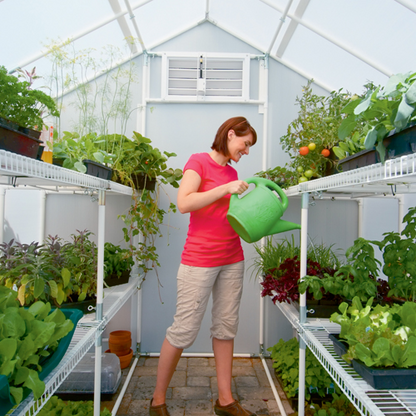 A smiling woman holding green watering can inside a solexx 8' X 8' Garden master small greenhouse watering the plants